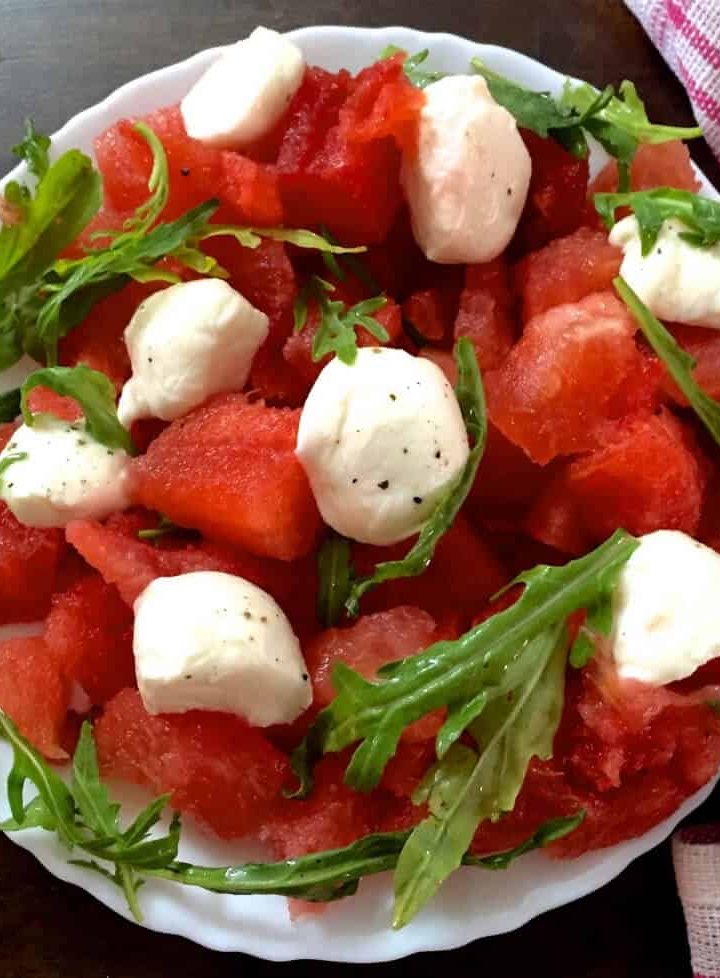 A white plate with a scalloped edge, filled with cubes of Watermelon and Halved Bocconcini cheese, with fresh arugula and pepper flakes for an easy cooling Watermelon Bocconcini Arugula Salad with Homemade Basil Oil