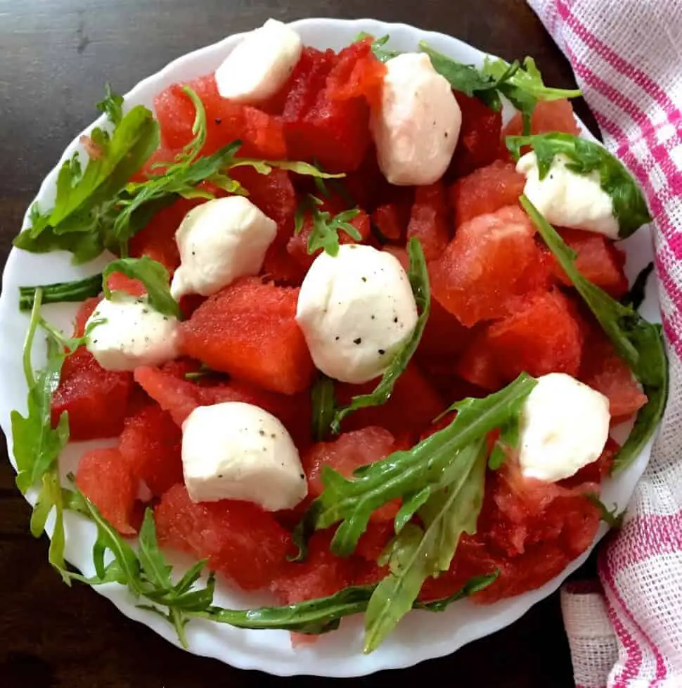 A white plate with a scalloped edge, filled with cubes of Watermelon and Halved Bocconcini cheese, with fresh arugula and pepper flakes for an easy cooling Watermelon Bocconcini Arugula Salad with Homemade Basil Oil