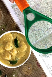 White bowl filled with a yellow creamy yogurt turmeric chickpea flour mixture cooked with colocasia root. An atlast with a map of the Indian State of Chattisgarh spread open on the right, with a magnifying glass highlighting the region
