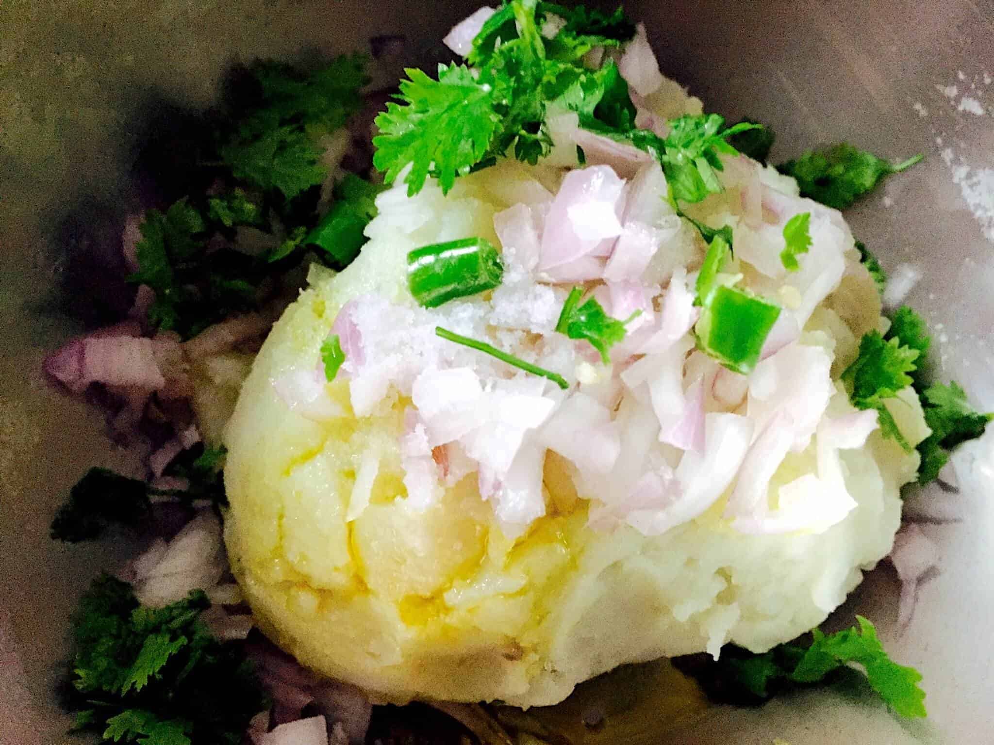 A large scoop of mashed potatoes, yellow on the left with mustard oil. Fresh coriander leaves and slices of green chili and onion and a scatter of salt are heaped up on the potoatoes. Some coriander leaves and onions scattered around