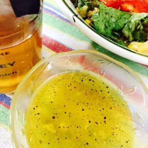 A bowl of yellow coloured fresh garlicky honey lemon salad dressing. A bottle of dark honey on the left and part of a leafy salad on the right, all on a striped rainbow coloured background