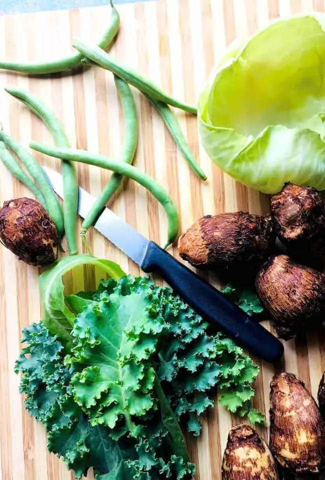 Ingredients for making Naga Hinkejvu or Colocasia Beans Greens Sabzi. Colocasia roots, Fresh greens, green beans and cabbage on a striped bamboo board with a black handled knife ready to slice the vegetables