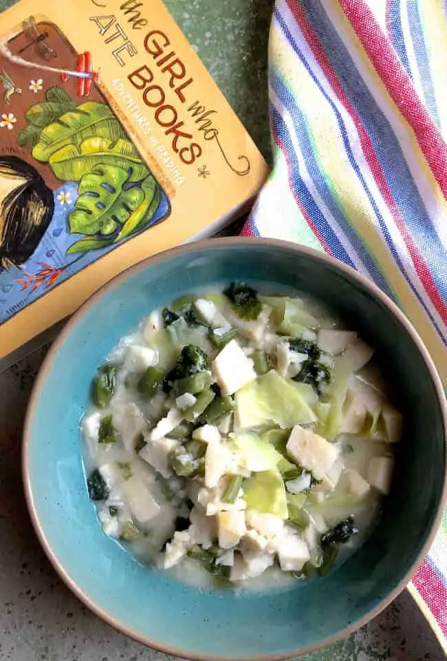 A sea green bowl edged with brown, with a whtie and green stew like mix of boiled colocasia slices, cabbage, beans and greens. Framed by a mustard book on the left (The Girl Who Ate Books) and a rainbow striped napkin on the top right