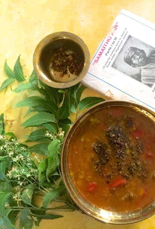 A brass bowl with brownish fluid Rasam, tempering of mustard, Fried neem flowers and cooked red tomato seen on top of the rasam. A bunch of fresh green neem leaves and flowers to the left and a brass bowl of fried neem flowers above along with a traditiona cookbook on Tamil Cuisine top right, all on a yellow background