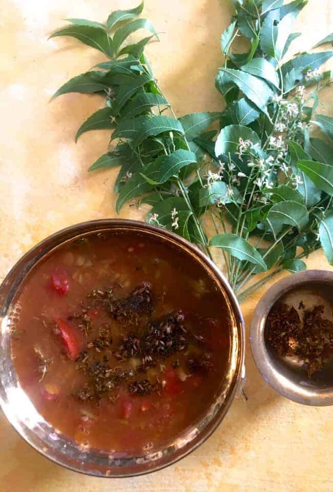 A bunch of fresh green neem leaves and flowers on a pale yellow background, with a braww bowl of brown coloured rasam, a fluid spicy tamarind dish. Red tomato slices, tempering of mustard seeds and neem flowers in ghee, seen on top of the rasam. A small brass bowl of fried neem flowerst o the right