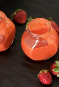 Two little jars of orangey pink strawberry poppyseed dressing, with strawberries strewn around them, all on a black background