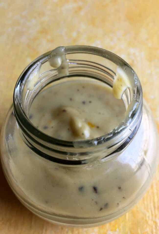 A round glass jar with a pale creamy yellow coloured tahini dressing with citrus and honey, flecked with black pepper powder, on a pale yellow background