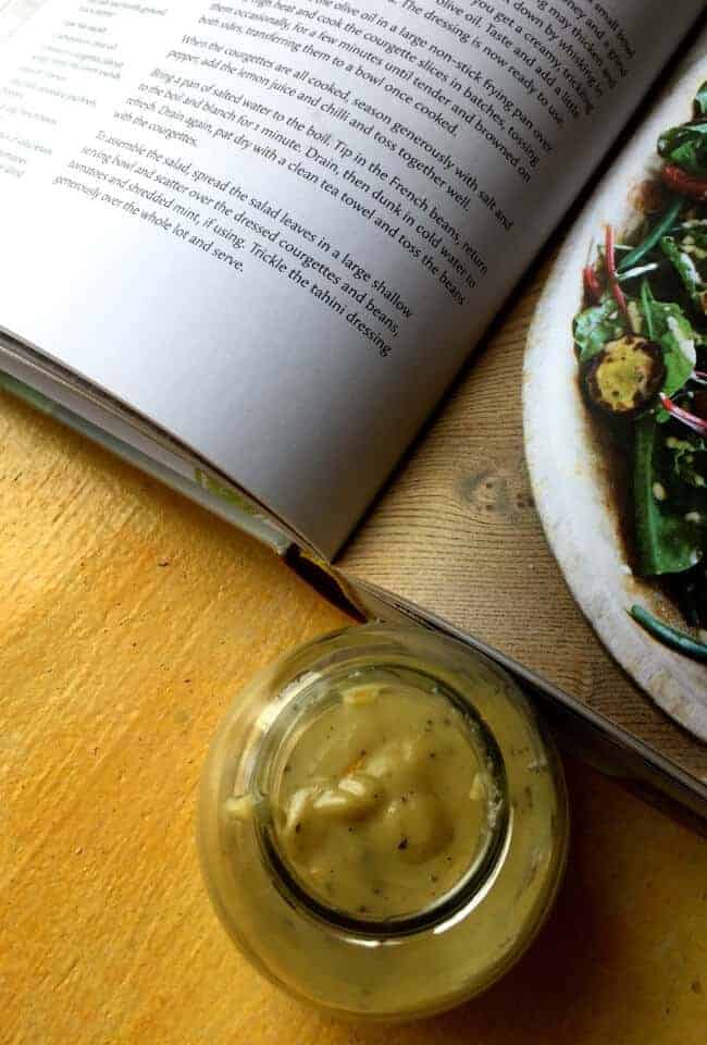 A small jar of creamy yellow salad dressing of tahini and citrus juices.A large cook book open beside, with part of a recipe for the salad dressing, and a part of a green salad on the facing page