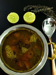 A bowl of dark fluid lemon thyme rasam with tomato, thyme leaves and mustard tempering seen on the surface. Two lemon halves above along with twigs of fresh thyme. A brass spoon to the right