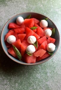 Bright red watermelon cubes and white spheres of mini bocconcini cheese in a blue green bowl edged with brown, on a green background. This Watermelon Bocconcini salad is gluten free and garnished with arugula leaves and basil