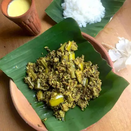 Green chunks of mixed vegetable curry in mustard paste and coconut, on a green piece of banana leaf resting on a clay plate. A clay tumbler in the bakcground with yellow mango lassi, white cooked rice on a banana leaf and a white hibiscus flower complete the presentation