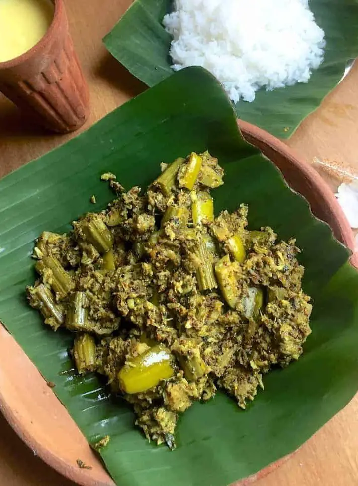 Green chunks of mixed vegetable curry in mustard paste and coconut, on a green piece of banana leaf resting on a clay plate. A clay tumbler in the bakcground with yellow mango lassi, white cooked rice on a banana leaf and a white hibiscus flower complete the presentation