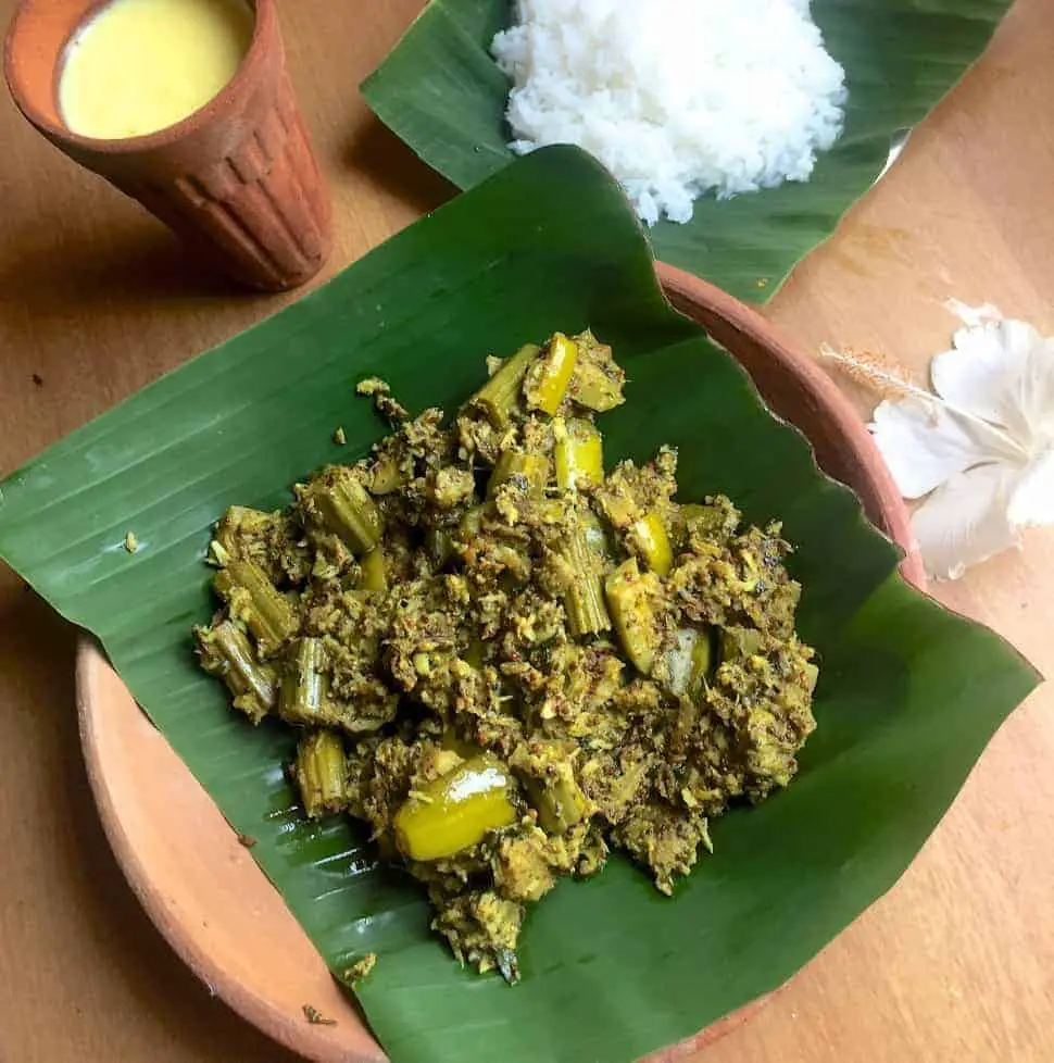 Green chunks of mixed vegetable curry in mustard paste and coconut, on a green piece of banana leaf resting on a clay plate. A clay tumbler in the bakcground with yellow mango lassi, white cooked rice on a banana leaf and a white hibiscus flower complete the presentation