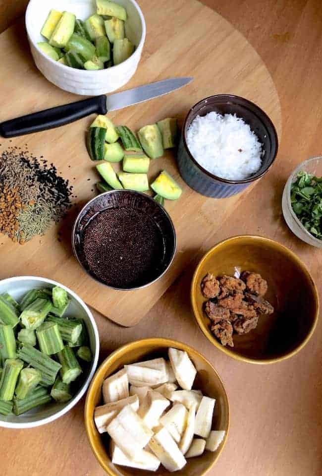 Ingredients for making the Traditional Besar or Besara - indigenous vegetables - slices of raw banana, parwal/potola or pointed gourd, drumstick, the traditional Odisha spice mix panch phutana, mustard seeds soaked for making mustard paste, moringa leaves, coconut and urad dal badi each in small cups, a knife for slicing veggies, resting on a cutting board