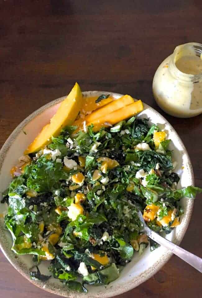 Massaged and marinated Kale mango salad with slices and cubes of ripe yellow mango and green kale, salad greens, yellow corn and white feta cheese. A small glass jar with cream coloured tahini dressing to the top right. All on a brown background