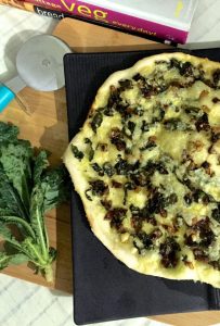 Thin crust Pizza on a black board, with kale and onion toppings. A bunch of kale to the left and a pizza cutter with a bright blue handle top left