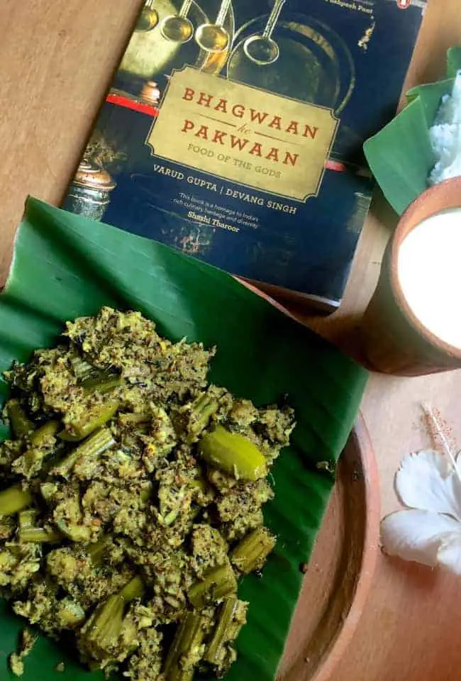 A black coverd book with red letters on a yellow background, 'Bhagwaan ke Pakwaan' and in the foreground a Besara vegetable curry on a green banana leaf on a clay plate, a white hibiscuis flower, cooked rice on another banana leaf and clay glass with yellow mango lassi