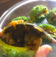 A baby bitter gourd/ karela held by the fingers while a stainless steel spoon of stuffing is being filled in. Other stuffed baby karela in the background