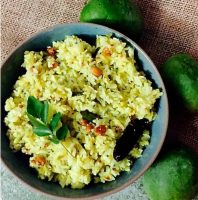 Ovehead view of a sea green bowl with yellow coloured raw mango rice resting against a piece of sackcloth with 3 green raw mangoes to the right. Mango rice is tempered with curry leaves, dry red chili and gram lentils