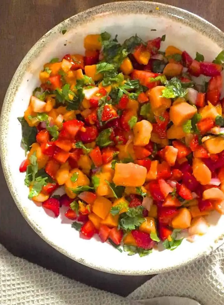 Overhead shot of a white bowl with yellow ripe mango slices, red strawberry slices, green basil and coriander leaves. A jar with yellow honey lemon salad dressing is on the left and a pale creamy grey napkin at the bottom of the image