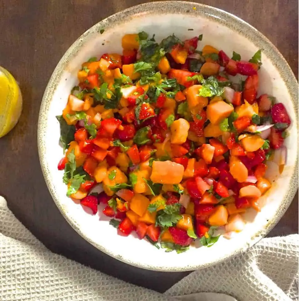 Overhead shot of a white bowl with yellow ripe mango slices, red strawberry slices, green basil and coriander leaves. A jar with yellow honey lemon salad dressing is on the left and a pale creamy grey napkin at the bottom of the image