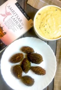 Overhead image of a white china plate with brown baby bitter gourd or karela, after stuffing and frying. Top left to the plate, a pale pink covered book called ' Amritsar to Lahore' and on the right. a bowl of yellow yogurt based Kadhi