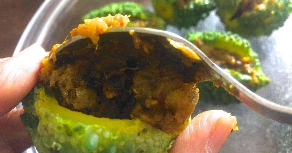 A bright green bitter gourd being stuffed with a brown spicy mango and onion filling using a stainless steel spoon. Other stuffed bitter gourds in the background, ready to be fried