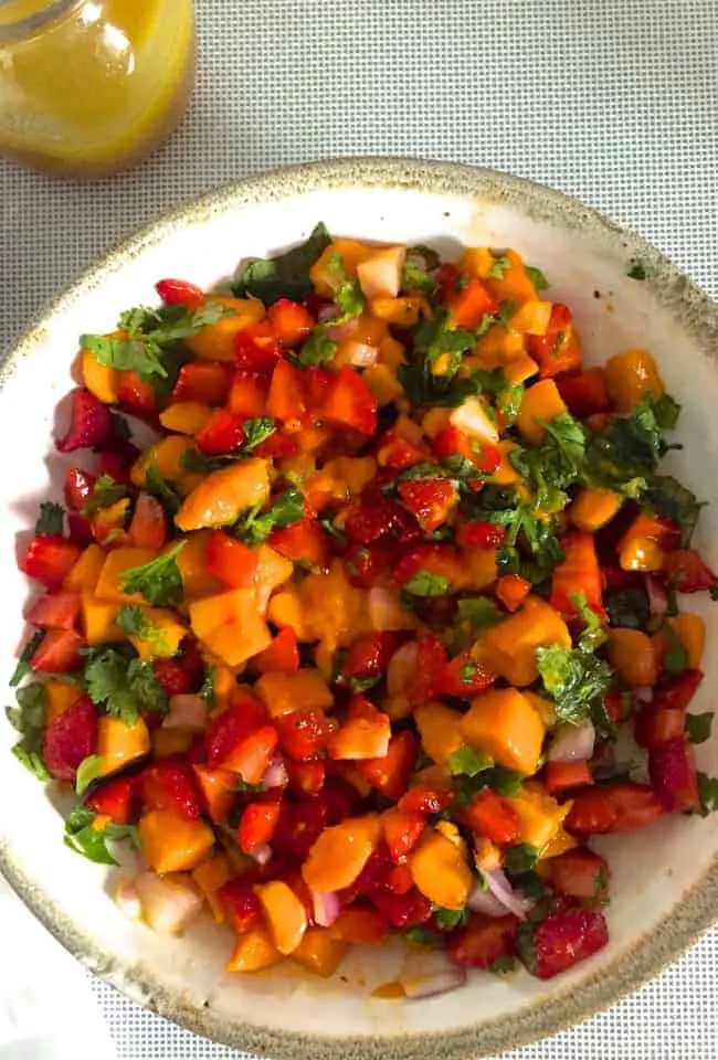 A white bowl seen from overhead, with bright coloured slices of orange yellow mango, red strawberry and flecks of green basil and coriander leaves 