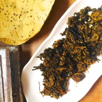 A plate of brown fried dry vegetables, Ker Sangri (berries and beans) and lotus stem slices on a long white plate with scalloped edges. A silver edged box and roasted pappad in the background