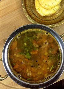 A brass bowl of Pineapple rasam garnished with curry leaves and small chunks of pineapple. A brass plate with slices of pineapple seen in the background, all on a wooden background