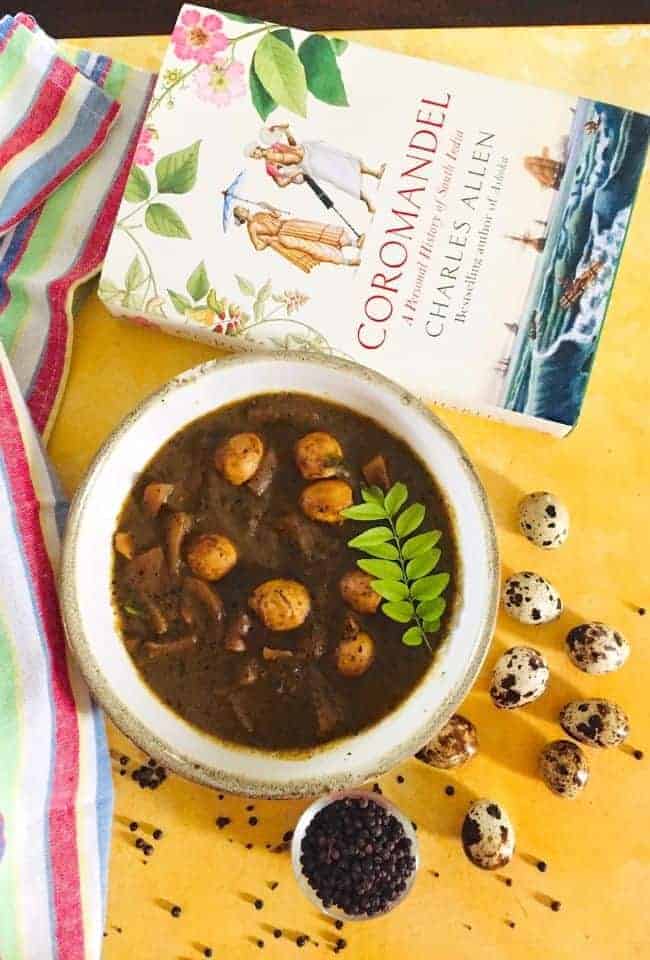 A white pottery bowl of quail eggs and slices of potato in a brown coloured pepper tamarind based gravy with a sprig of curry leaves on the surface. A striped red, green white napkin lies on the left and a white covered book Coramandel, in the background. Some quail eggs and scattered peppercorns in the foreground