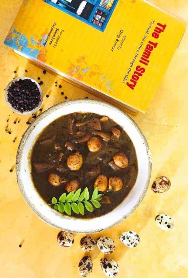 A bowl of dark brown coloured pepper gravy with quail eggs and potatoes and a sprig of curry leaves on top.All on a yellow background with a yellow coloured book called The Tamil Story (in red letters) in the background. A pot of black peppercorns and scatter pepper are behind the bowl. Some quail eggs are in the foreground