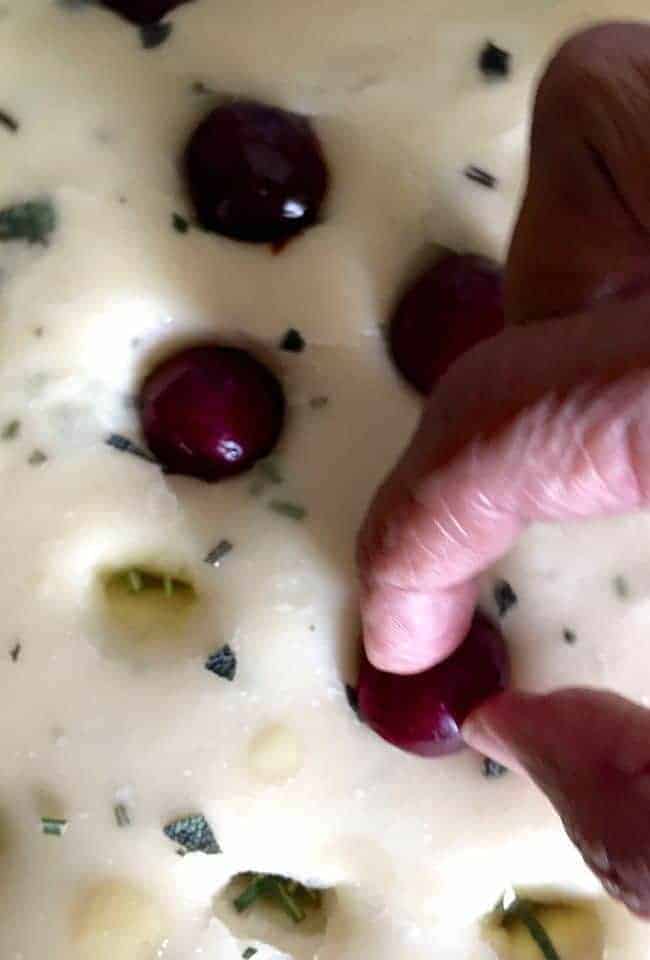 Cherry halves being placed in indentations on soft focaccia dough, filled with olive oil. Fingers seen placing the cherry half into the indentation