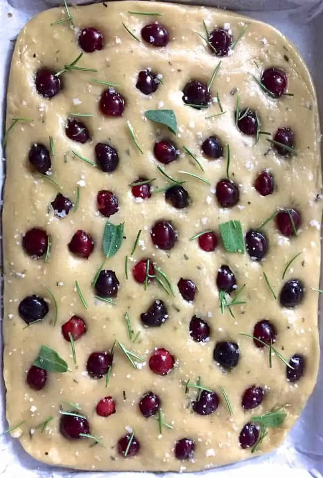 Cherry Focaccia ready to go into the oven, studded with cherry halves and with sea salt, olive oil and a garnish of rosemary and sage leaves