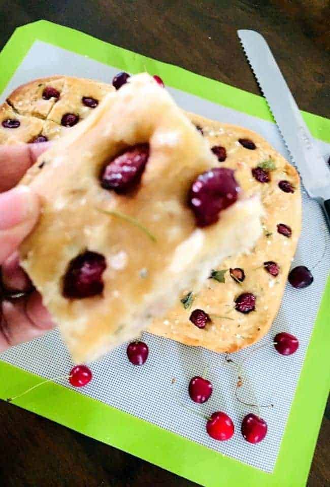 A slice of creamy yellow Focaccia bread studded with cherry halves, and a freshly baked focaccia seen below on green edged mat, with cherries scattered on it