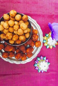 Plate of brownish round vella seedai with a bowl of lighter coloured uppu seedai on top, on a petunia pink silk fabric. Small silver lamps and a purple flower on the side