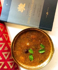 Lentil and spice coconut paste cooked in tamarind extract. Sambar in a brass bowl with a few small green curry leaves and the tops of small Madras onions floating on the surface. A grey and black Tamil classic book in the background and a red cloth with gold print on the left. The whole on a white background