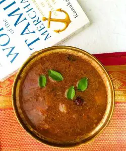 A large brass bowl filed with brown arachuvitta sambar, with little green curry leaves and the tops of small onions on the surface. Resting on an orange and red rich silk zari saree. A white book with green titles in the background and all on a white background