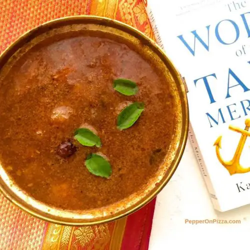 Brown coloured sambar in a brass bowl, with green curry leaves and pale white and red small onions floating on top. The bowl rests on an orange and red zari silk saree and has a white book with blue letters on Tamil merchants, on the right. All on a white background