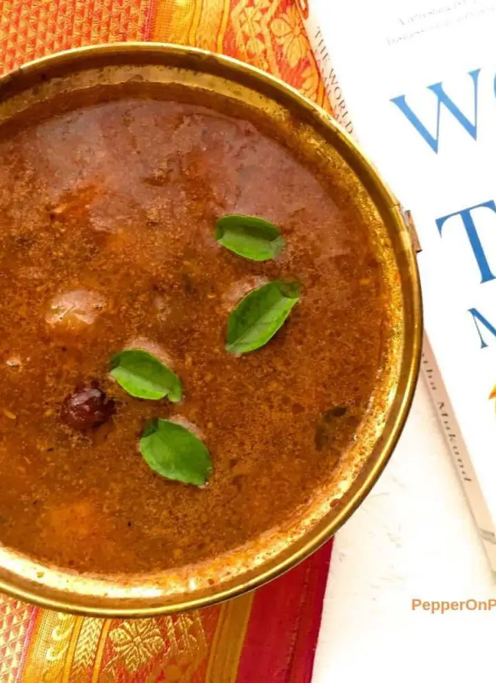 Brown coloured sambar in a brass bowl, with green curry leaves and pale white and red small onions floating on top. The bowl rests on an orange and red zari silk saree and has a white book with blue letters on Tamil merchants, on the right. All on a white background