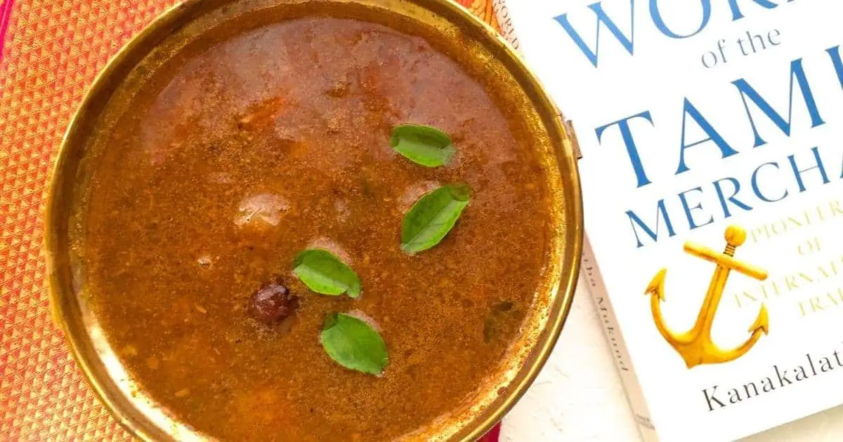 A brass bowl of arachuvitta sambar from Tamil cuisine. Brown tamarind gravy with little sambar onions and curry leaves floating on top. Part of a white book with blue headings seen on the right, the whole on a white background