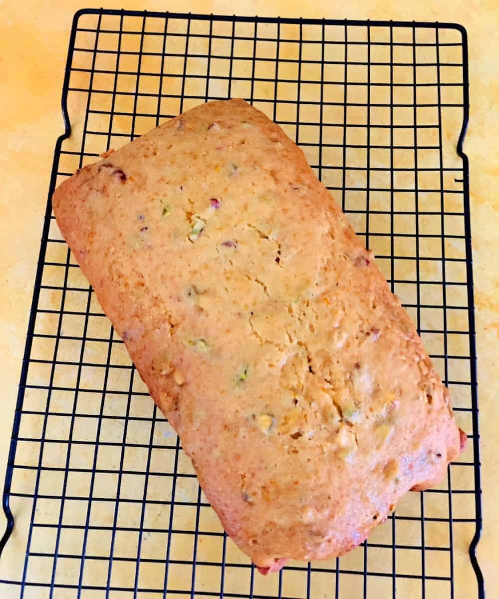 Loaf of golden orange honey tea cake studded with pistachio slices, on a wire rack, all on a yellow background board