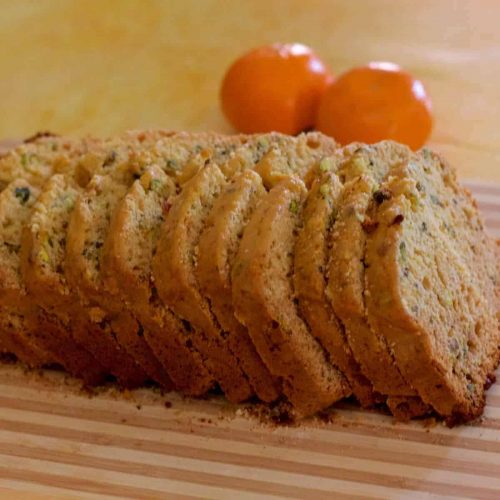 Slices of pale golden orange honey pistachio cake, on a striped board, with 2 bright oranges in the background, all on a pale yellow backdrop