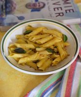 Yellow coloured penne pasta in pumpkin sauce flecked with red chili flakes and garnished with basil leaves, in a white pasta plate rimmed with green. A cook book with white blue and brown cover image in the background, and red green yellow striped napkin on the right. All of this on a yellow background