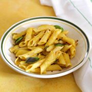Penne pasta in a yellow pumpkin sauce with green basil leaves and red chili flakes, on a white pasta plate edged with green, on a yellow background. A white napkin with thin green stripes on the right