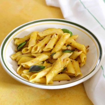 Penne pasta in a yellow pumpkin sauce with green basil leaves and red chili flakes, on a white pasta plate edged with green, on a yellow background. A white napkin with thin green stripes on the right