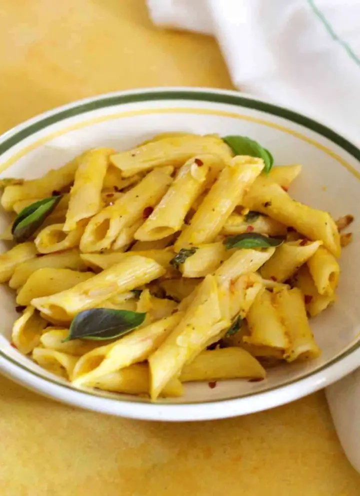 Penne pasta in a yellow pumpkin sauce with green basil leaves and red chili flakes, on a white pasta plate edged with green, on a yellow background. A white napkin with thin green stripes on the right