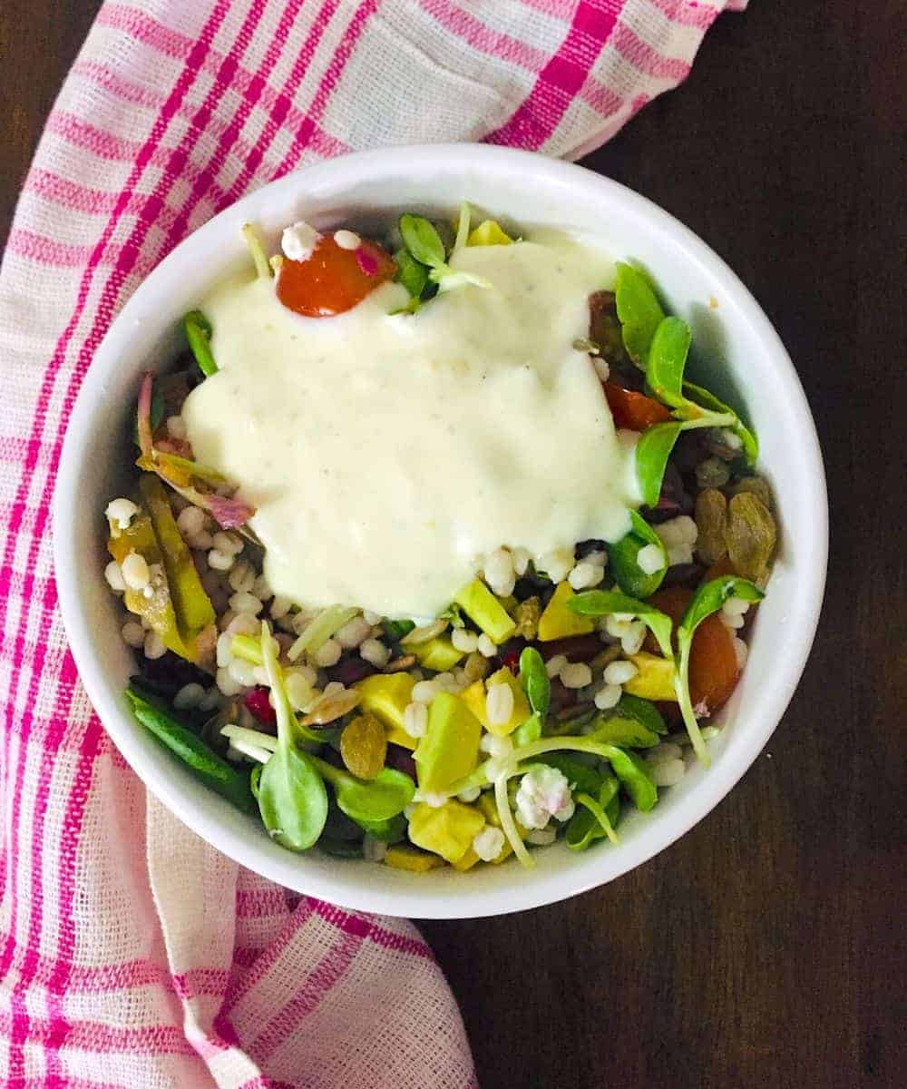 Pearl Barley Tomato Avocado Salad with microgreens and with hung curd dressing spread on top on one side, in a bowl and with a pink and white checked napkin to the left, all on a dark brown background