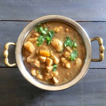 Thiruvathirai Ezhukari kootu, a stew from 7 vegetables in a brown tamraind gravy, garnished with cilantro, in a 2 handled brass bowl, on a gray wooden background