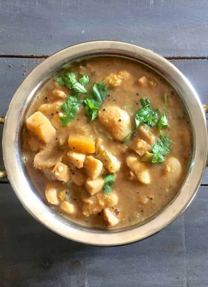 Thiruvathirai Ezhukari kootu, a stew from 7 vegetables in a brown tamraind gravy, garnished with cilantro, in a 2 handled brass bowl, on a gray wooden background